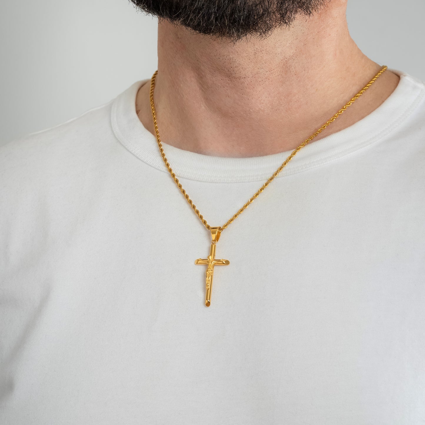 A male model in a white t-shirt wearing a Jesus Crucifix Minimalist Cross Gold Pendant with a 3mm Gold Rope chain 22 inches. Close-up image of the non-tarnish trending religious men's necklace.