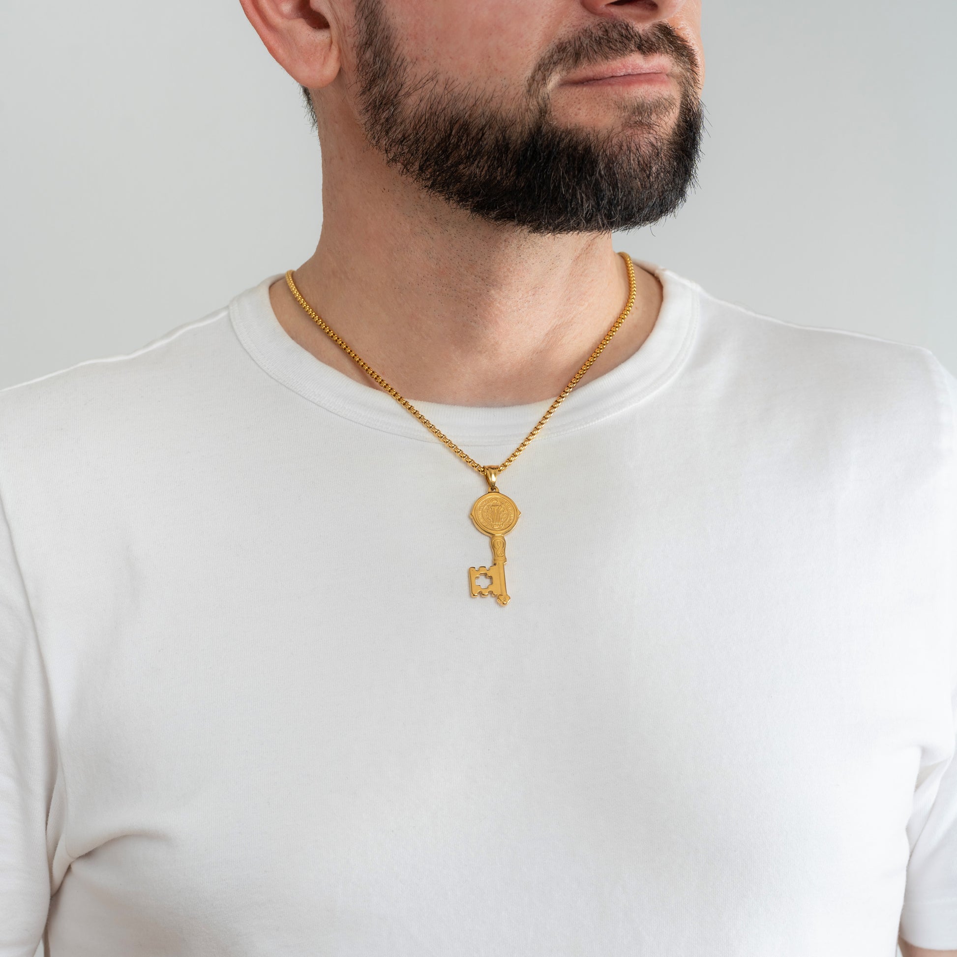 A male model in a white t-shirt wearing a St. Benedict Key Gold Pendant with a 3mm Gold Round Box link chain 22 inches.