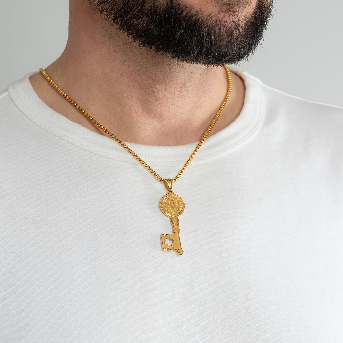 A male model in a white t-shirt wearing a St. Benedict Key Gold Pendant with a 3mm Gold Round Box link chain 22 inches. Close-up image of the non-tarnish trending men's necklace.
