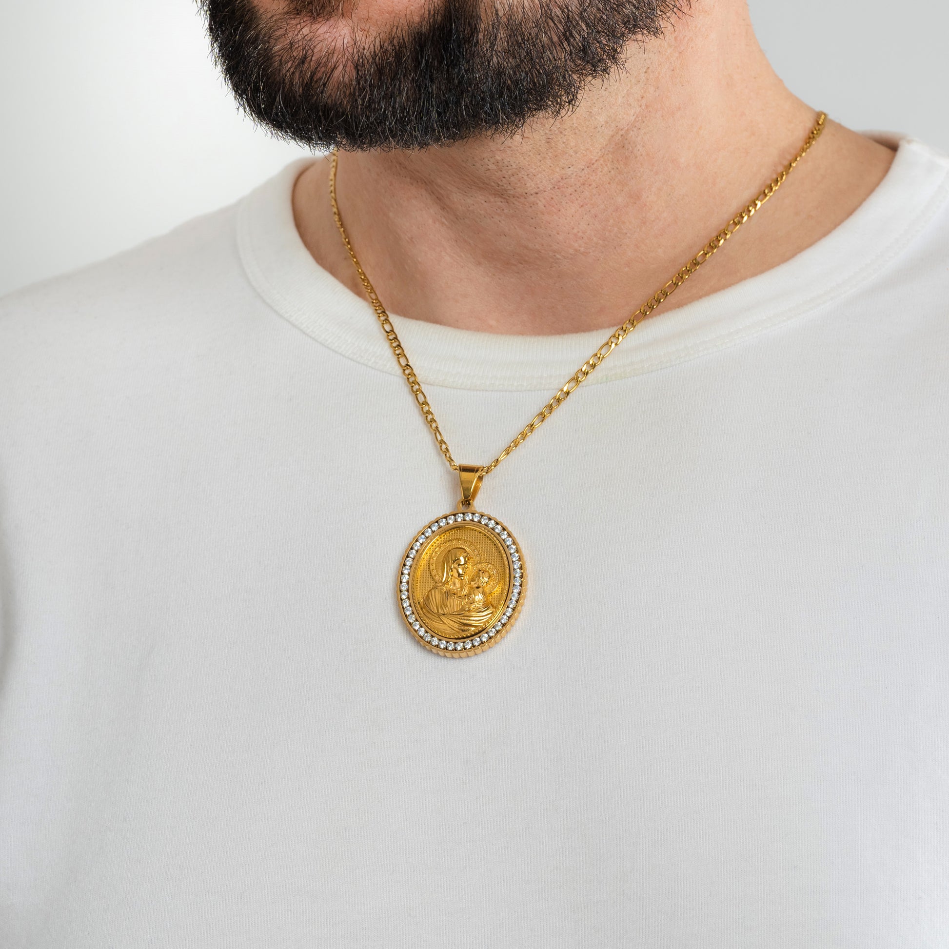 A male model in a white t-shirt wearing a Mary, Mother of Jesus Gold Pendant with a 3mm Figaro Gold chain 22 inches. Close-up image of the non-tarnish religious men's necklace with Cubic Zirconia inlays.