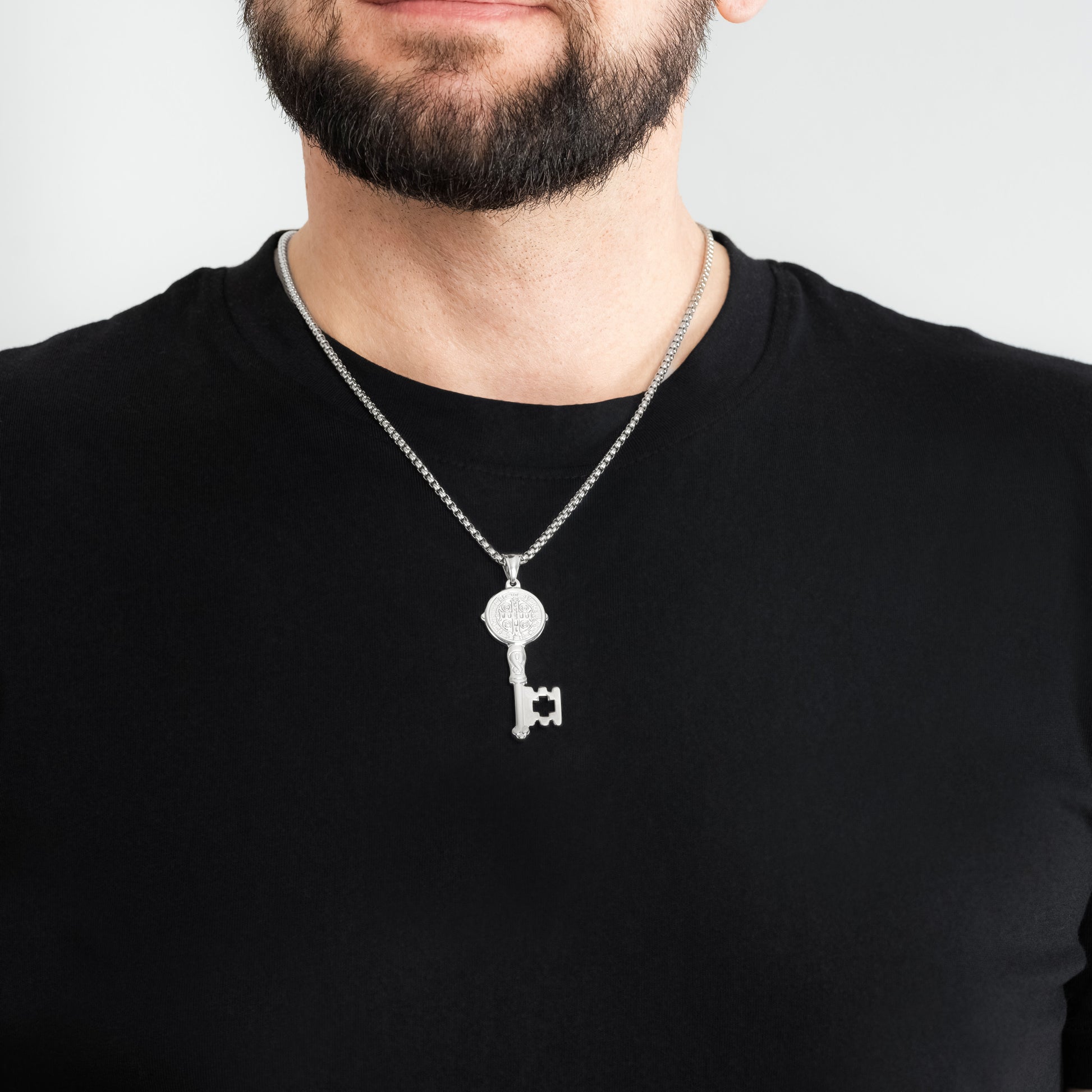 A male model in a black t-shirt wearing a St. Benedict Key Silver Pendant with a 3mm Silver Round Box link chain 22 inches.
