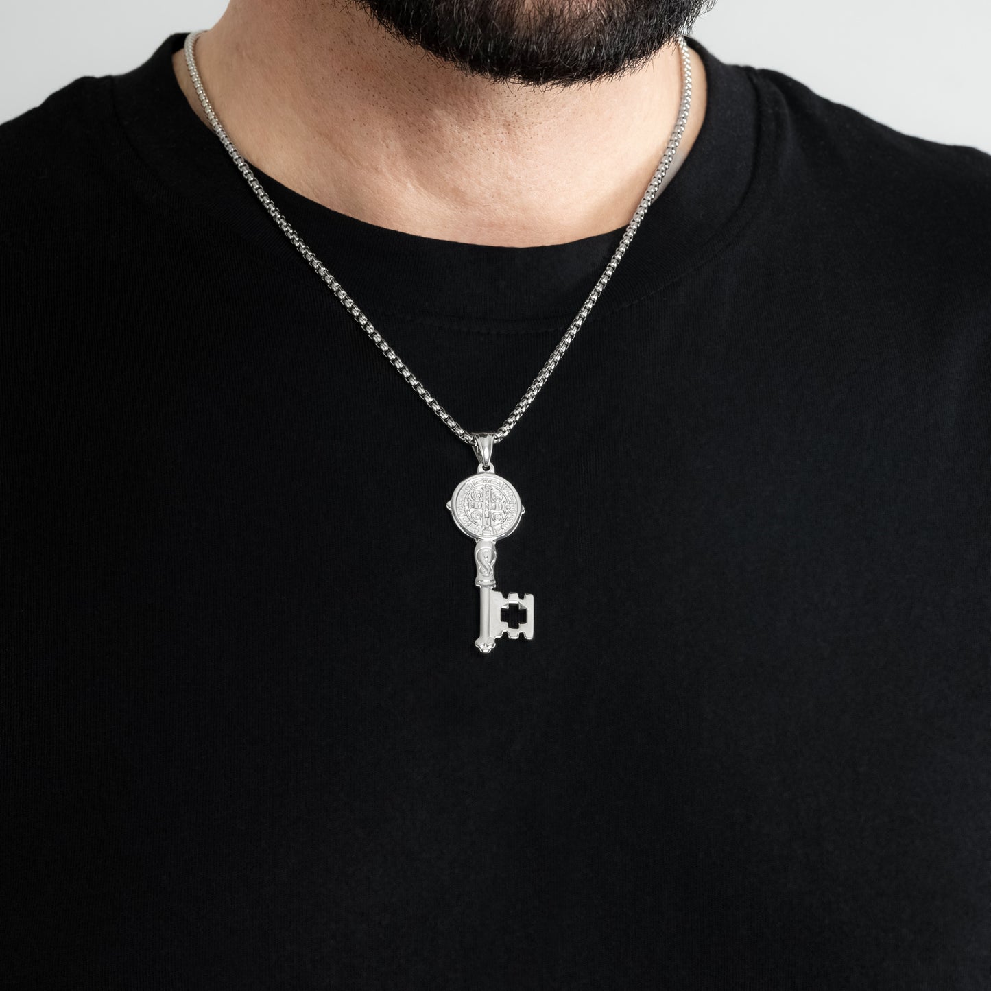 A male model in a black t-shirt wearing a St. Benedict Key Silver Pendant with a 3mm Silver Round Box link chain 22 inches. Close-up image of the non-tarnish meaningful men's necklace.