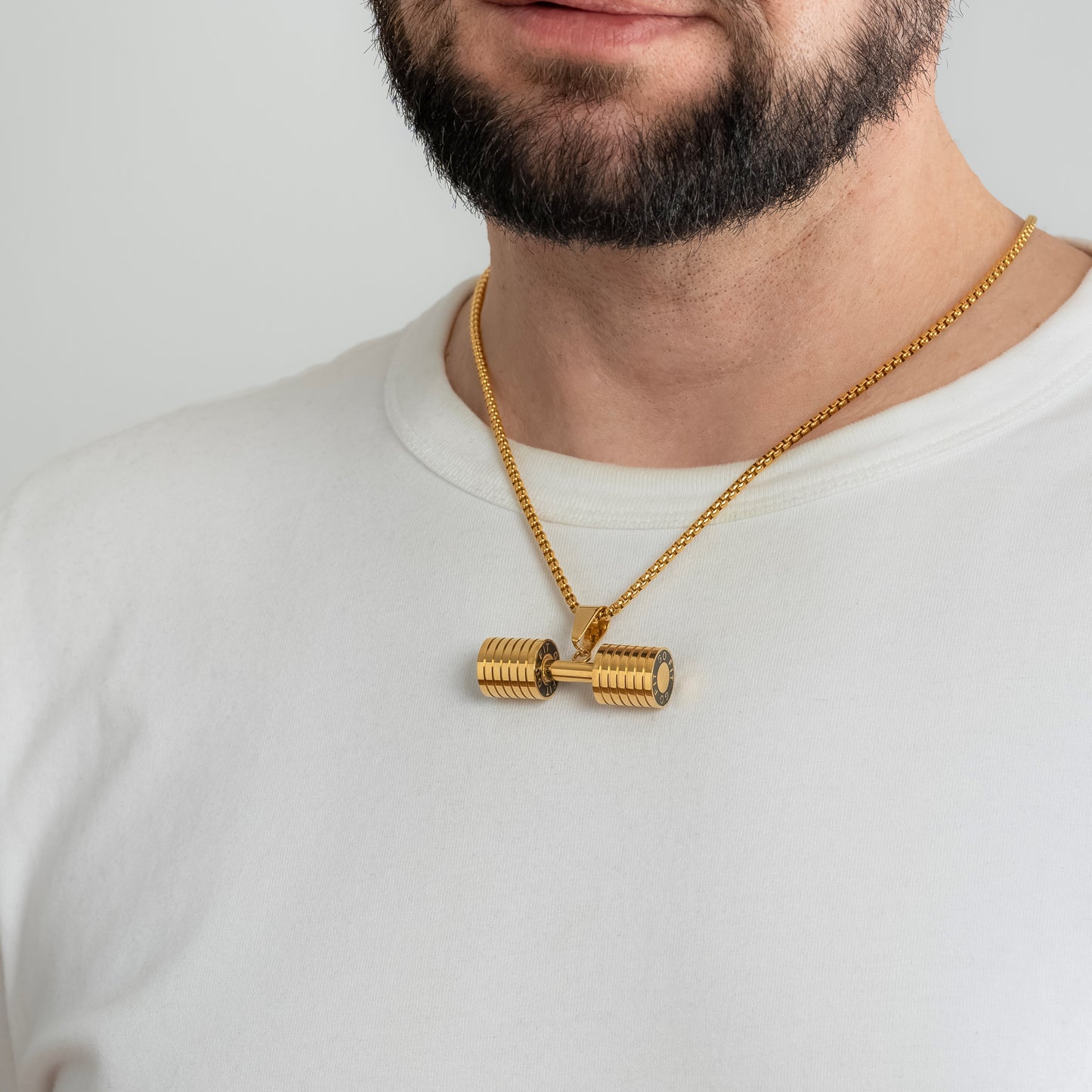 A male model in a white t-shirt wearing a Dumbbell Fitness Gold Pendant with a 3mm Round Box link Gold chain 22 inches. Close-up image of the tarnish-free, sweatproof and waterproof motivational men's necklace.